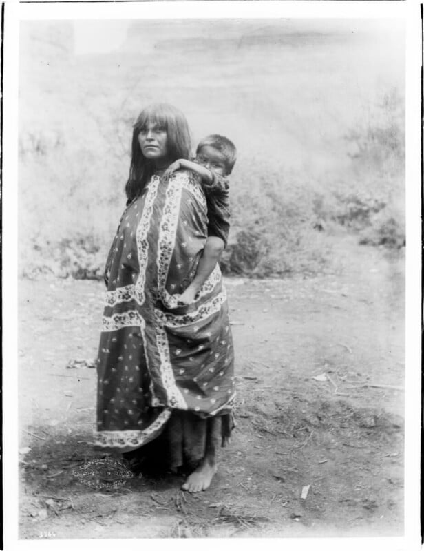 A barefoot woman stands outdoors, wrapped in a patterned blanket, carrying a young child on her back. The child looks over her shoulder, both have straight dark hair. The background is natural and slightly blurred.