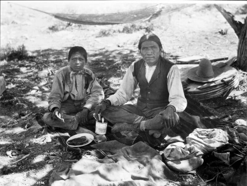 Two Indigenous men sit on the ground outdoors among blankets and supplies, with food dishes in front of them. Trees and a hammock are in the background, with a hat resting nearby.
