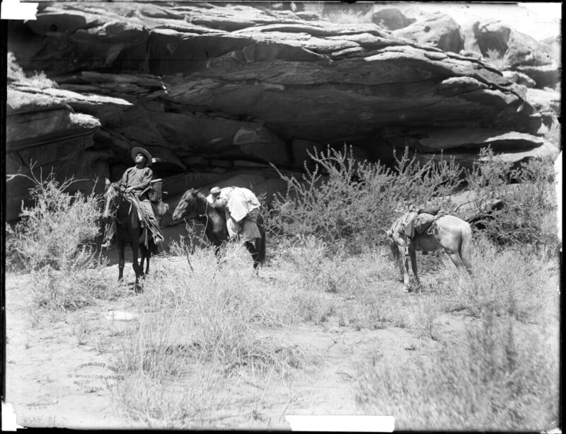 A person in traditional clothing sits on a horse next to two saddled horses in a dry, rocky landscape with large boulders and sparse vegetation.