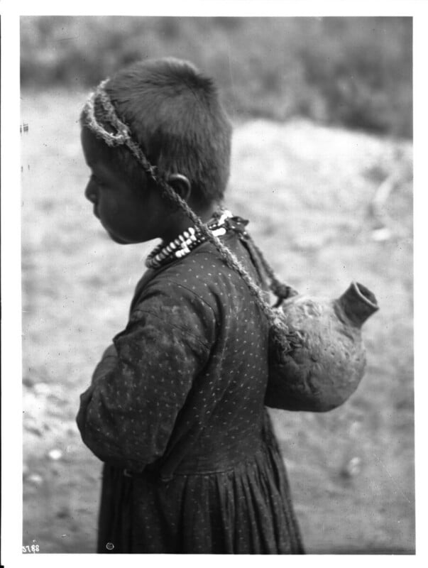 A young child with short hair, wearing a patterned dress and beaded necklace, stands in profile carrying a clay pot tied with string on their back. The background is blurred and outdoors.