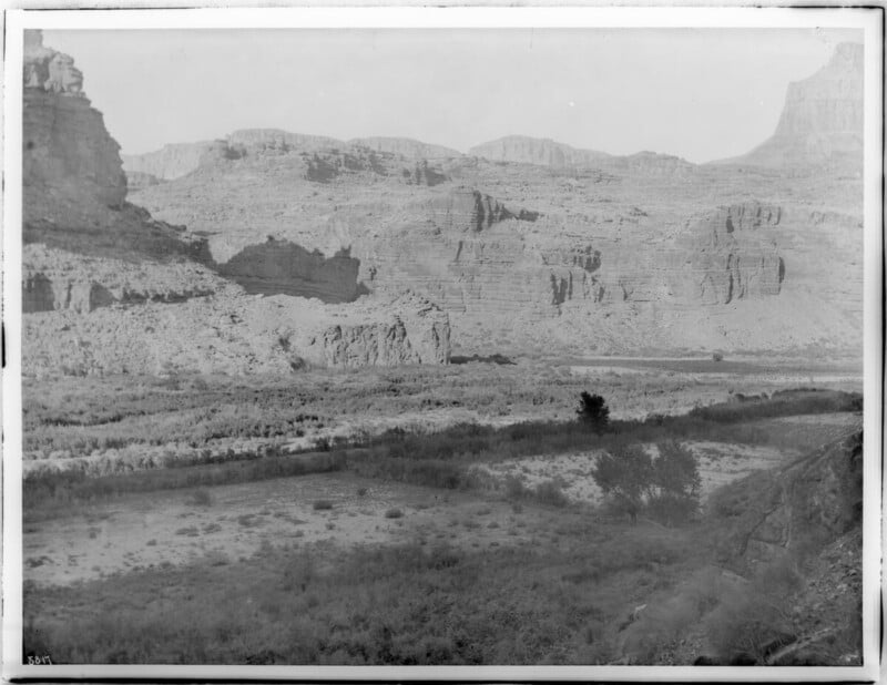 Black and white photo of a vast canyon landscape with layered rock formations, a river running through the middle, and sparse vegetation in the foreground. The scene appears rugged and arid.