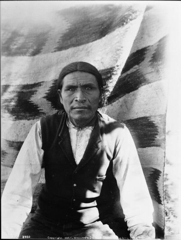 A Native American man with long hair, wearing a vest over a light shirt, sits in front of a patterned blanket hanging as a backdrop. He looks directly at the camera with a serious expression.
