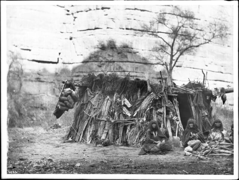 Three people sit in front of a small, rustic shelter made from branches and leaves, set against a rocky cliff. A tree stands nearby, and various objects are scattered on the ground around them.