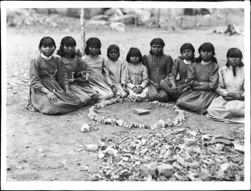 A group of young Native American girls sit in a circle on the ground outside, surrounding a ring of stones with a single stone in the center. They wear long dresses and have straight, dark hair with bangs.