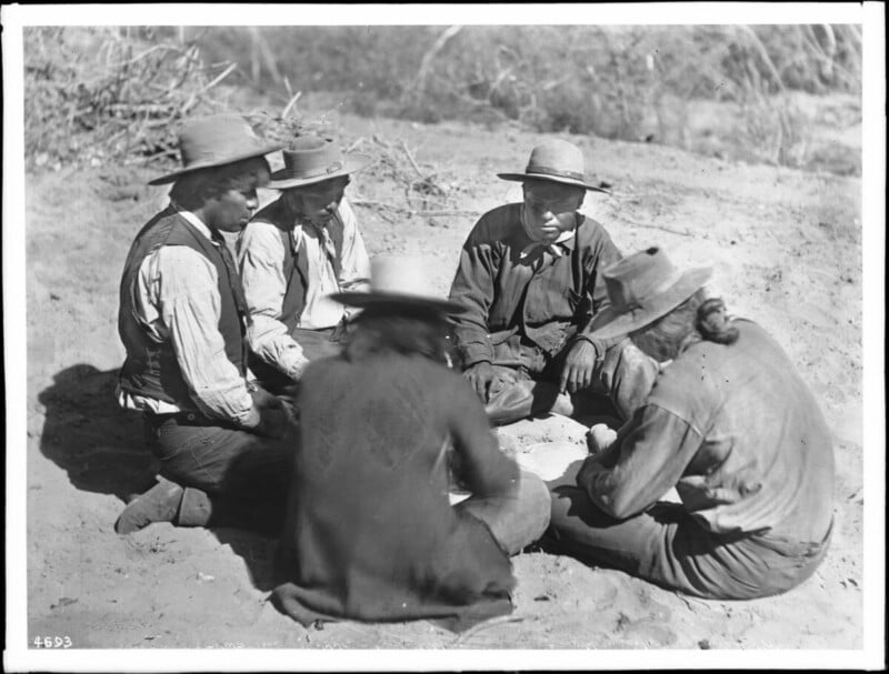 Five people wearing hats and long-sleeved clothing sit in a circle on sandy ground outdoors, appearing to engage in conversation or an activity together. Bushes and dry landscape are visible in the background.