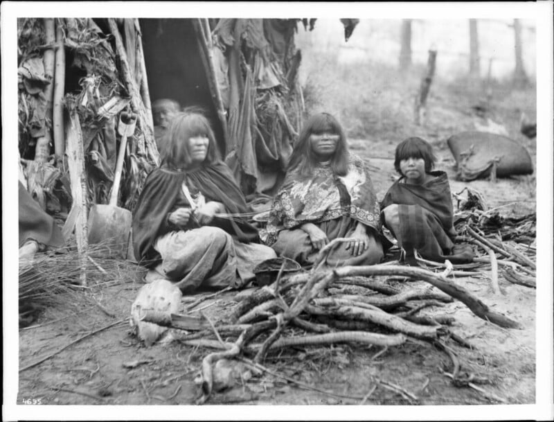Three Indigenous women and a child sit on the ground near a pile of sticks and branches, in front of a structure made from natural materials. The scene appears outdoors with a blurred natural background.