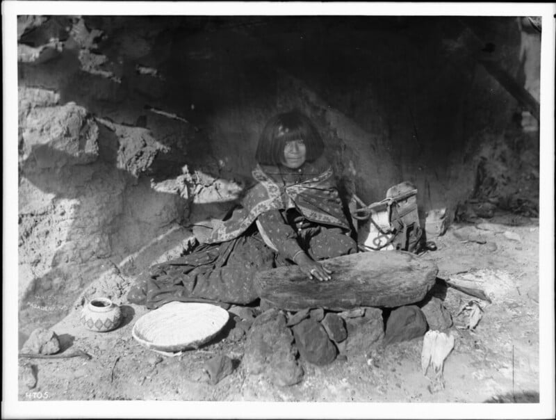 A young Indigenous girl sits on the ground in front of a stone structure, wrapped in a patterned blanket, with baskets and traditional tools arranged around her. She extends her hand over a flat stone in a sunlit outdoor setting.