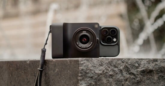 A smartphone with a Leica camera attachment and lens sits upright on a stone surface outdoors, with a blurred background of water fountains.