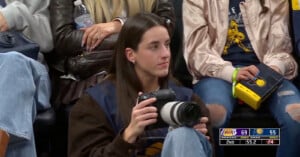 A woman sits in the crowd at a basketball game holding a camera. The scoreboard on the screen shows Lakers 69, Pacers 55, with 55.2 seconds left in the 2nd quarter. Other spectators are seated around her.
