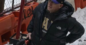 A man in a black puffer jacket and blue baseball cap stands outdoors on snow-covered ground near orange barricades and a metal fence with graffiti. He is holding a black bag.