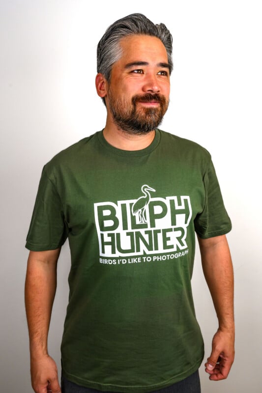 A man with gray hair and a beard is wearing a green t-shirt that says "BIRPH HUNTER" with a bird graphic and the text "BIRDS I'D LIKE TO PHOTOGRAPH." He is smiling and standing against a white background.