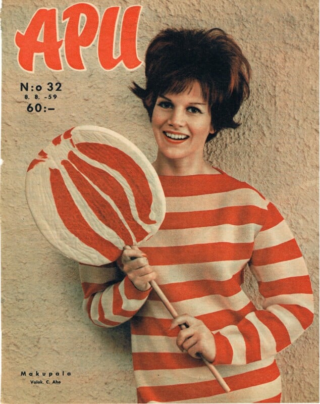 A smiling woman in a red-and-white striped top holds a large, matching red-and-white striped lollipop. She stands against a textured beige wall with "APU" magazine text and issue details above her.