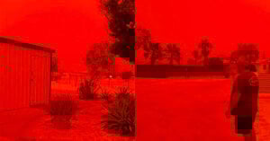 A landscape is bathed in an intense red-orange hue, with buildings, trees, and a person walking outside. The sky appears completely red, likely due to a dust storm or severe atmospheric conditions.