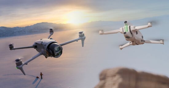 Two drones fly over a snowy landscape at sunset; mountains and a person standing alone on the snow are visible below, with the sun low near the horizon.