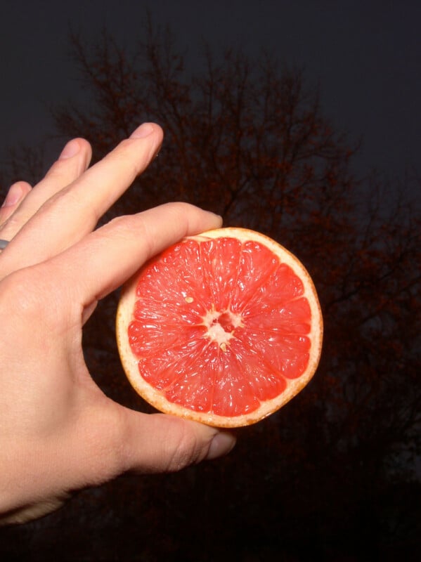 A hand holds a half of a juicy, red grapefruit against a dark outdoor background with the faint outline of tree branches.