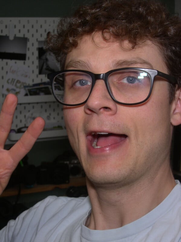 A person with curly hair and glasses smiles at the camera, holding up a peace sign with their hand. The background features shelves with cameras and various items.