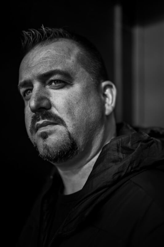A man with short hair and a goatee looks seriously at the camera in a black and white portrait. He is wearing a dark jacket and standing against a dark, blurred background.