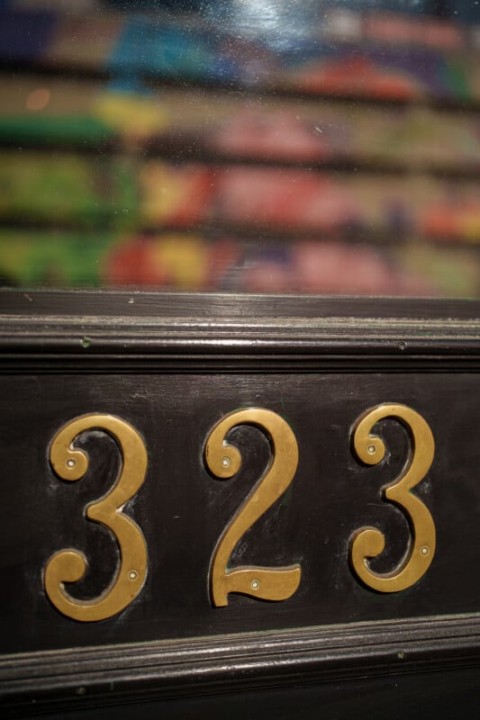 Gold numbers "323" are displayed on a dark wooden surface, with a slightly blurred background showing rows of colorful objects behind a glass window.