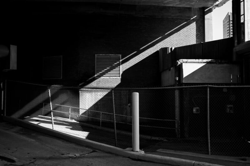 A black and white photo of an urban alleyway under an overpass, featuring a chain-link fence, brick walls with vents, and a diagonal beam of sunlight cutting through the shadows.