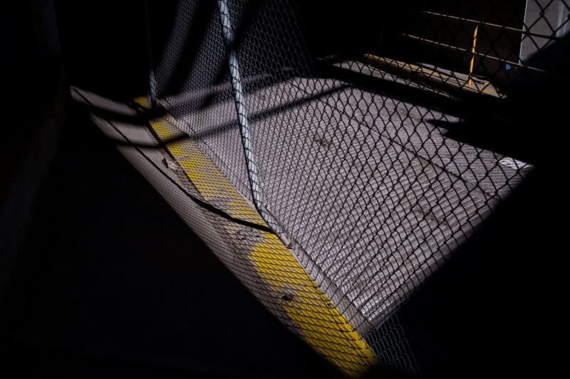 A chain-link fence casts shadows on pavement lit by strong sunlight, highlighting a bright yellow painted line on the ground. The scene appears to be part of an industrial or parking area.