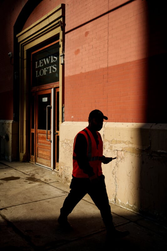 A person in a red safety vest and cap walks past the entrance to LEWIS LOFTS, with sunlight casting strong shadows on the building’s red brick and tan walls. The person appears to be looking at a phone.