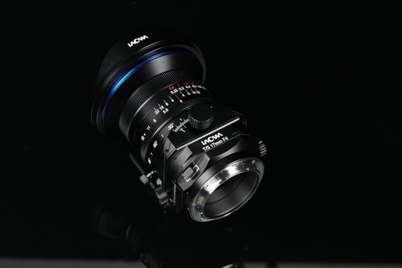 A Laowa camera lens with adjustment rings and detailed markings is displayed on a glossy black surface, reflecting some light.