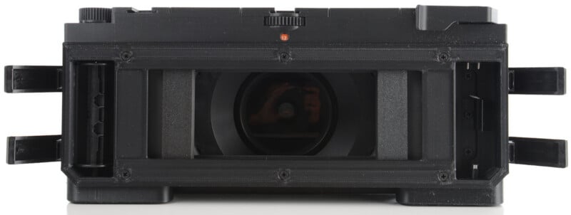 A close-up, front-facing view of a black rectangular camera film holder with side flaps open and the lens visible in the center.