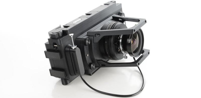A black, boxy large-format film camera with a wide lens and metal support brackets sits on a white surface, with a cable release attached to the lens.