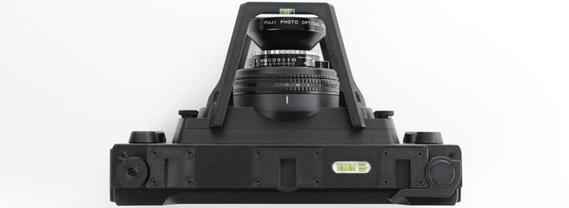 A black large format camera viewed from above, showing a wide-angle lens, a spirit level, various knobs, and adjustment mechanisms against a white background.