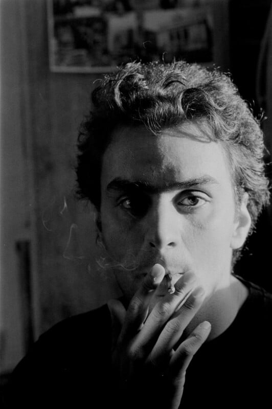 A person with curly hair smokes a cigarette, gazing intently at the camera. Light and shadow create dramatic contrast on their face. The background is blurry, with some photographs or posters visible on the wall.