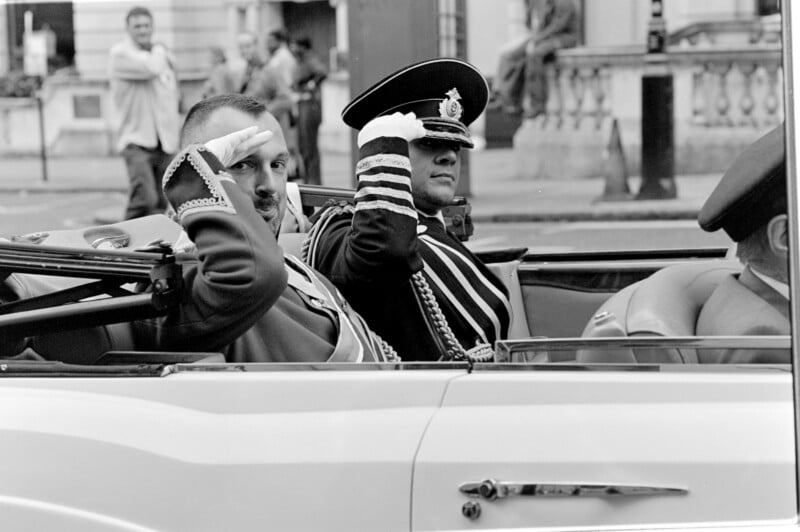 Two men in formal military uniforms and hats are sitting in a convertible car, saluting as they pass by. The photo is black and white, and people and buildings are visible in the background.