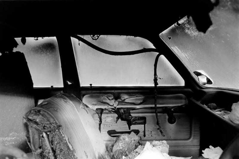 Interior of a severely burned car with melted upholstery, damaged seats, and soot covering the windows, showing extensive fire damage and charred debris inside the vehicle.