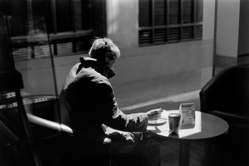 A person sits alone at a small round table by a window, eating and drinking. Sunlight streams in, casting shadows. The scene is in black and white, with an empty chair nearby and a coffee cup on the table.