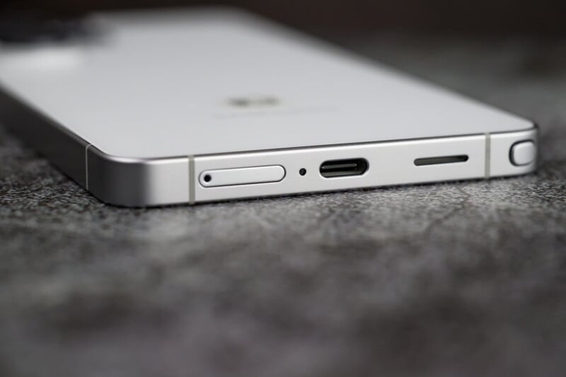 Close-up of the bottom edge of a white smartphone showing a USB-C charging port, speaker grille, microphone, and SIM card tray on a gray textured surface.