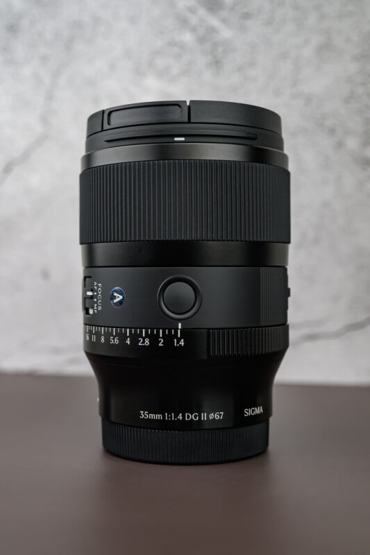 A Sigma 35mm f/1.4 DG DN Art camera lens stands upright on a brown surface with a light gray, textured background. The lens is black with visible markings and adjustment rings.
