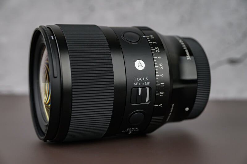 A close-up of a black camera lens with focus and aperture adjustment dials, labeled markings, and textured grip panels, placed on a smooth surface with a blurred gray background.