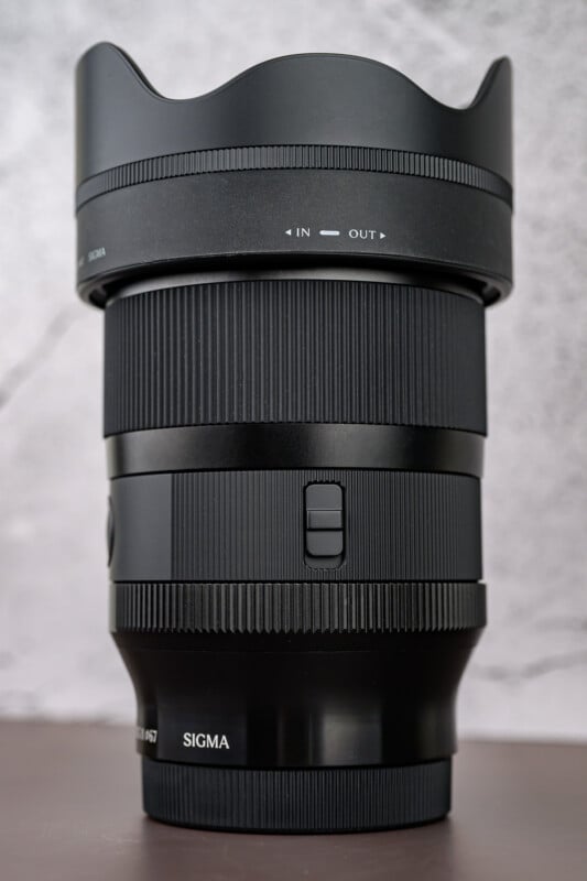 A close-up of a Sigma camera lens with a lens hood attached, placed upright on a smooth surface against a light, textured background. The lens features focus and zoom rings and labeled controls.