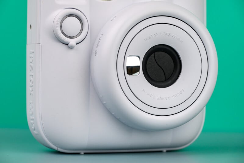 Primer plano de una cámara instantánea blanca con lente circular sobresaliente y visor pequeño sobre fondo cian sólido.