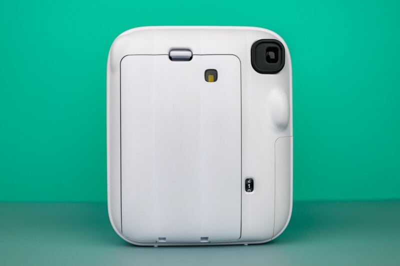 Vista posterior de una cámara instantánea cuadrada blanca que muestra el compartimento de la batería y el área de la lente sobre un fondo cian.