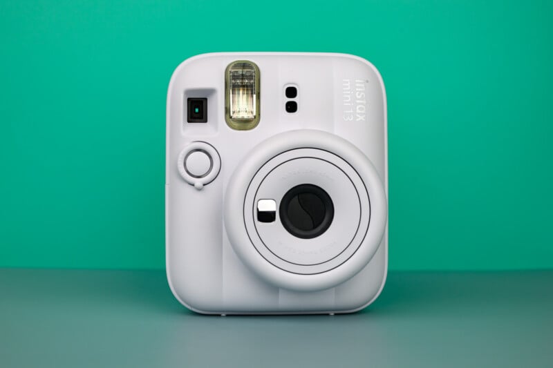 Una cámara instantánea Instax Mini 11 blanca colocada sobre una superficie plana con un fondo cian. La lente de la cámara y el flash son claramente visibles.