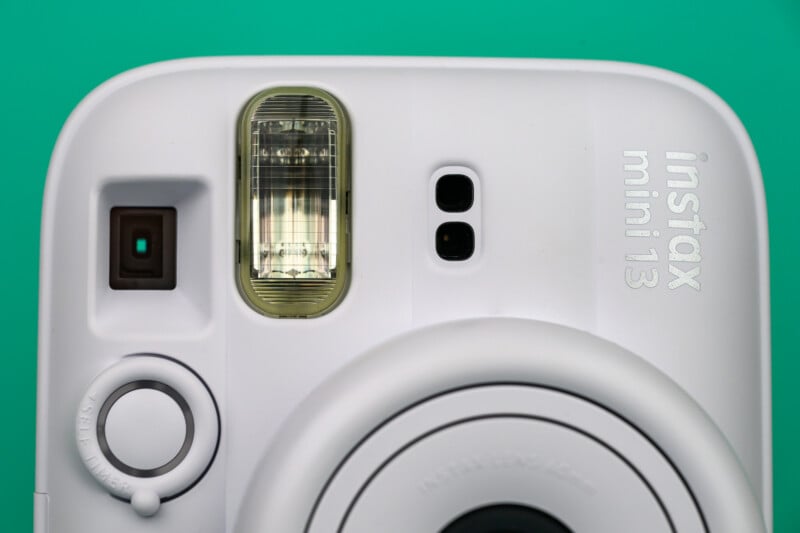 Primer plano del frente de una cámara instantánea Instax Mini 13 blanca con un fondo cian que muestra la lente, el flash y el logotipo.