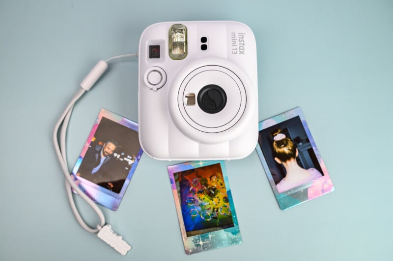 Una cámara Polaroid Instax Mini 11 blanca se encuentra sobre una superficie azul claro, rodeada por tres fotografías Polaroid reveladas que muestran una variedad de escenas coloridas, así como un hombre con un moño, visto desde atrás.