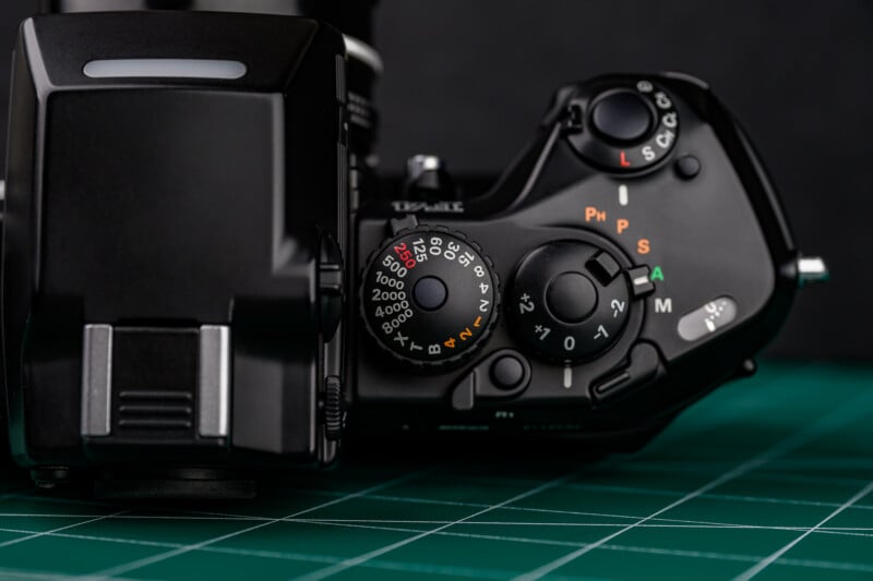 Close-up of a black digital camera's top controls, including mode dial, power switch, and shutter button, resting on a green cutting mat with black background.