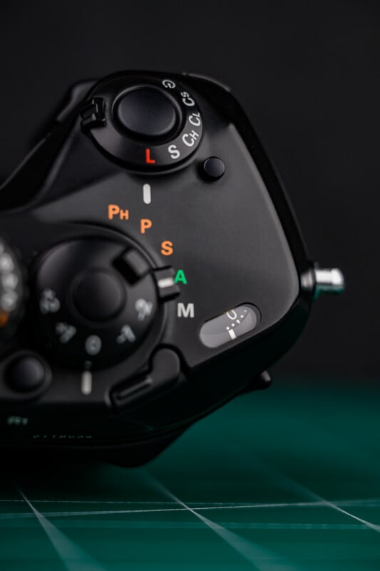 Close-up of a black camera mode dial with various settings labeled in white, orange, and green letters, positioned on a dark background with a green grid surface below.