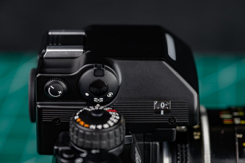 Close-up view of the top of a black analog camera showing dials, buttons, and a small display on a green cutting mat background. The camera's controls and texture are clearly visible.