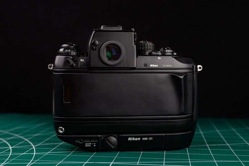 A Nikon film camera viewed from the back, featuring a black body, visible viewfinder, control dials, and attached MB-21 grip, set on a green cutting mat with a black background.