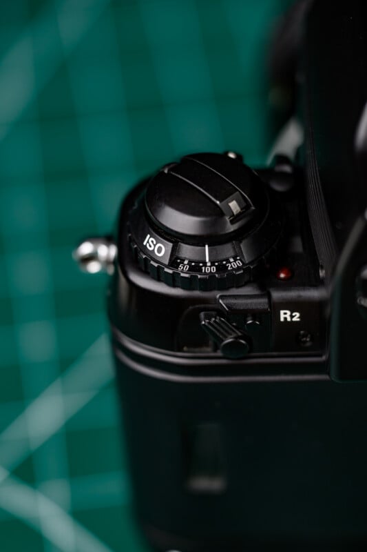 Close-up of a black DSLR camera’s ISO dial, set on a green cutting mat with a grid pattern. The dial displays ISO values from 25 to 200. The image focuses on the camera’s top-left corner.