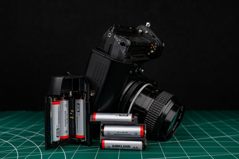 A film camera with its battery compartment open sits on a green cutting mat. Several AA batteries, some inside the compartment and some loose, are visible against a black background.