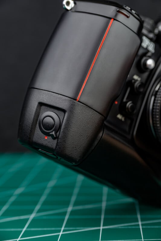 Close-up of a black camera body with a vertical grip and red accent line, placed on a green cutting mat with white grid lines. The grip features a black shutter button with a small red dot.