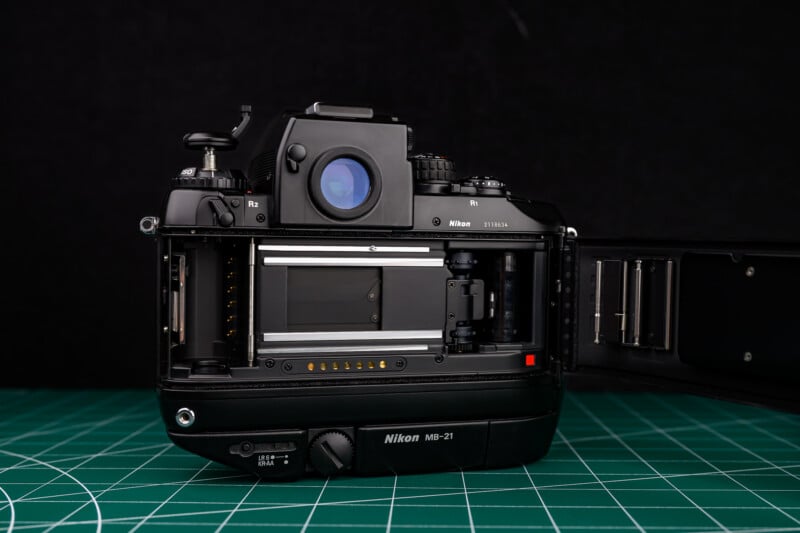 A Nikon film camera with its back cover open, revealing the film compartment and mechanisms, sits on a green cutting mat with a black background.